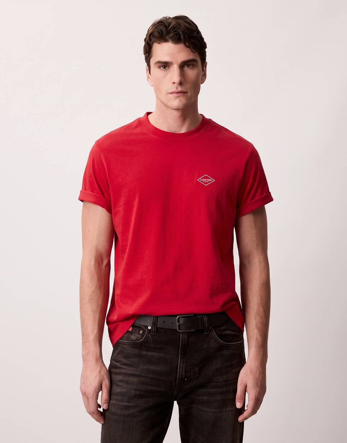 A male model stands, wearing a Calvin Klein 20s Matchbox Graphic Red T-Shirt paired with dark jeans. The red t-shirt