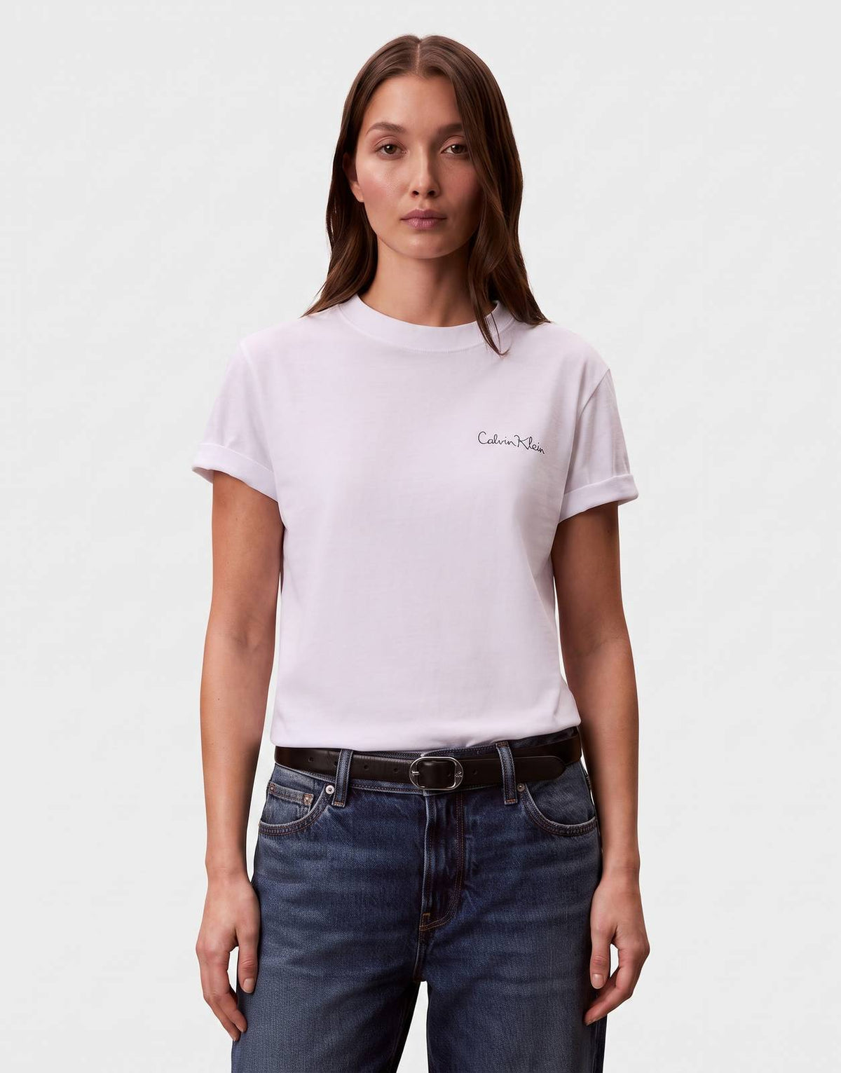 A woman models a Calvin Klein Classic Graphic White T-Shirt featuring a black handwritten logo, paired with dark wash jeans.