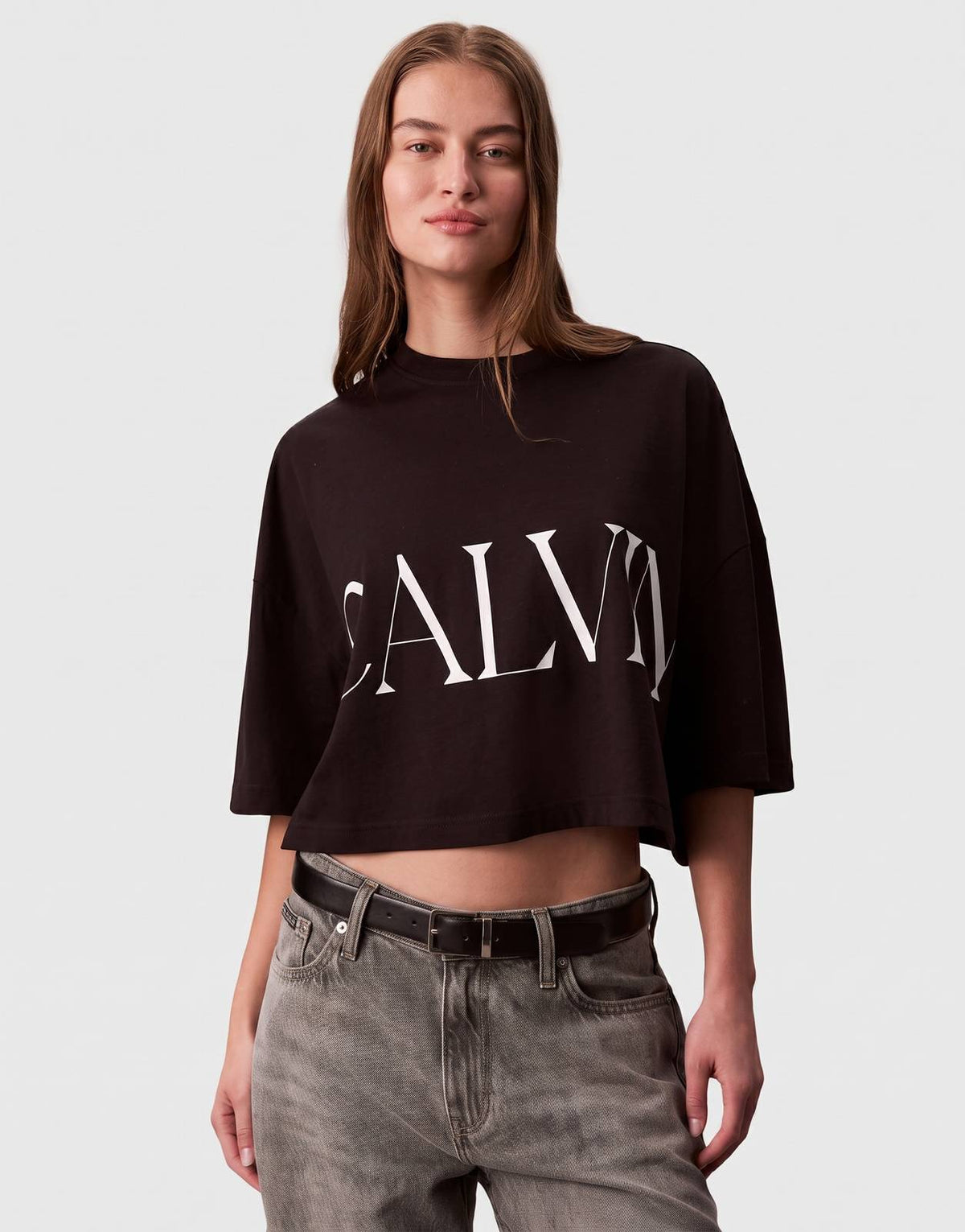 A female model showcases a black Calvin Klein Cropped Graphic T-Shirt with a white logo print on the front, captured in a