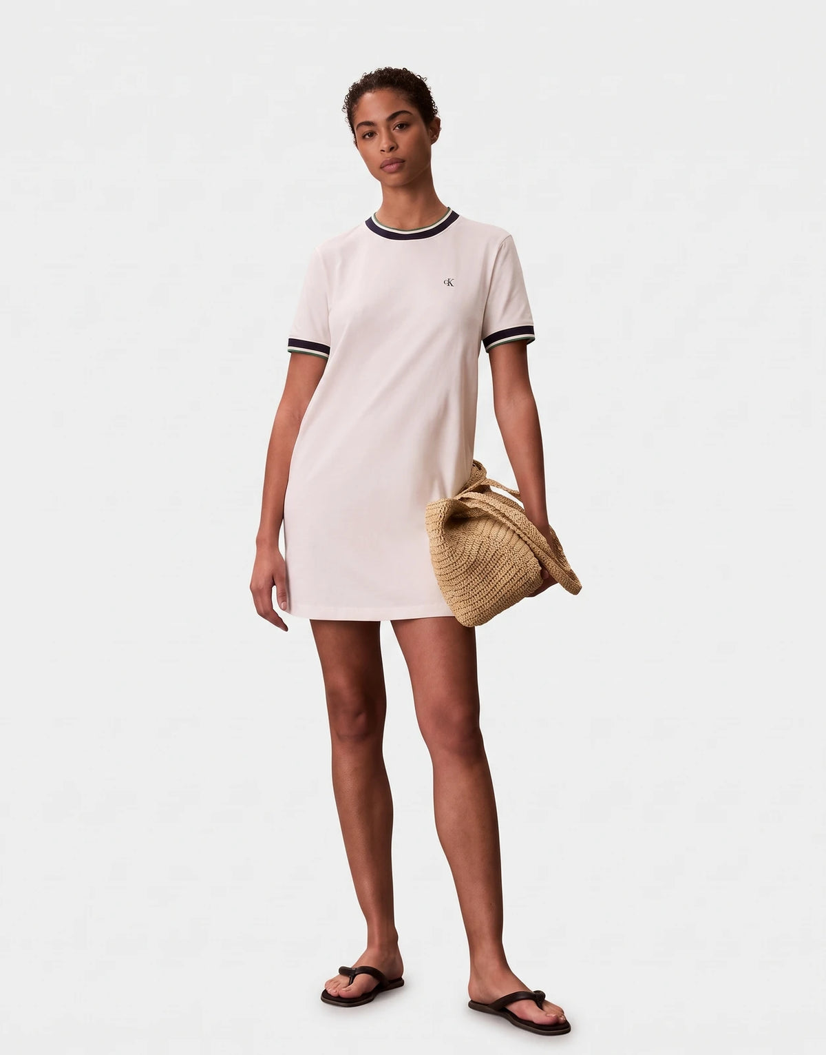 Full body shot of a female model wearing an off-white Calvin Klein cotton monogram stripe trim mini dress and holding a