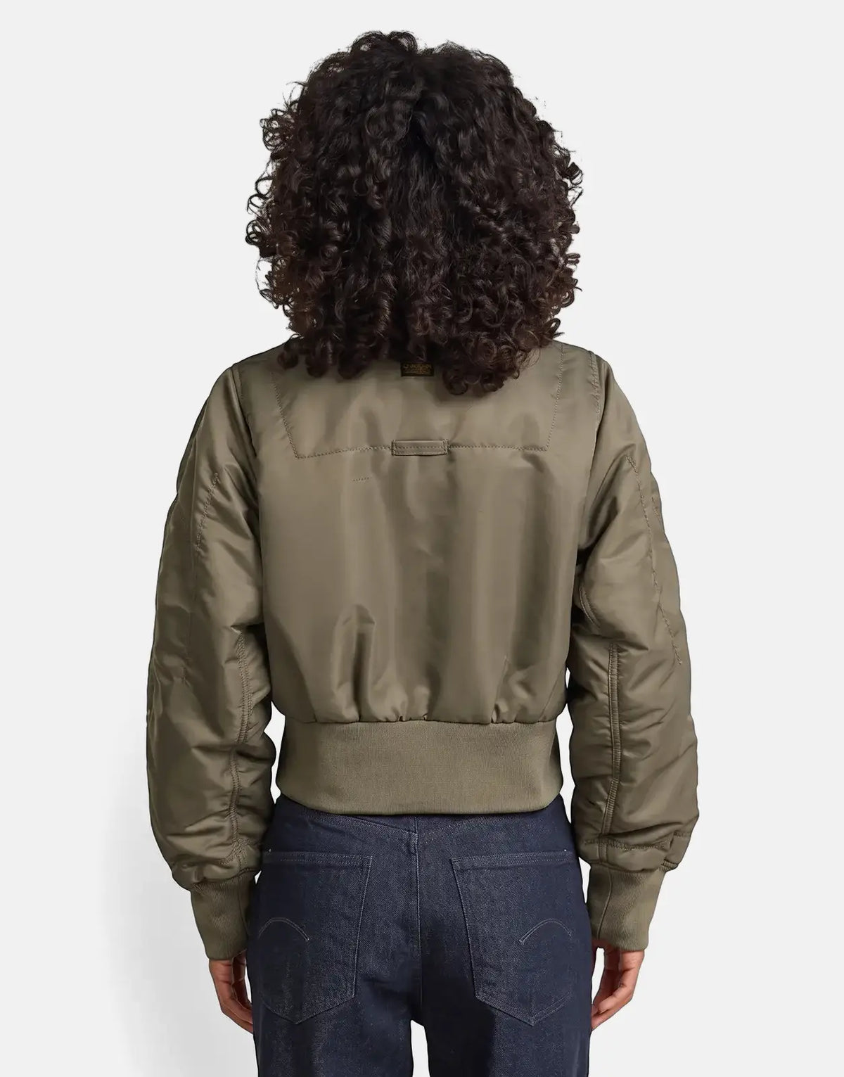 A woman from behind models an olive green G-Star RAW Bomber Turf jacket and dark jeans, showcasing the jacket's ribbed