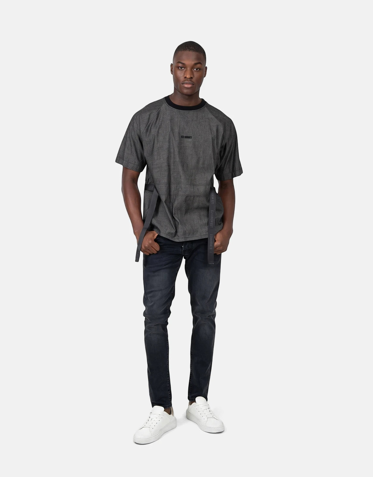 A male model wearing a grey Les Hommes t-shirt with black belt-like straps, black jeans, and white sneakers, standing