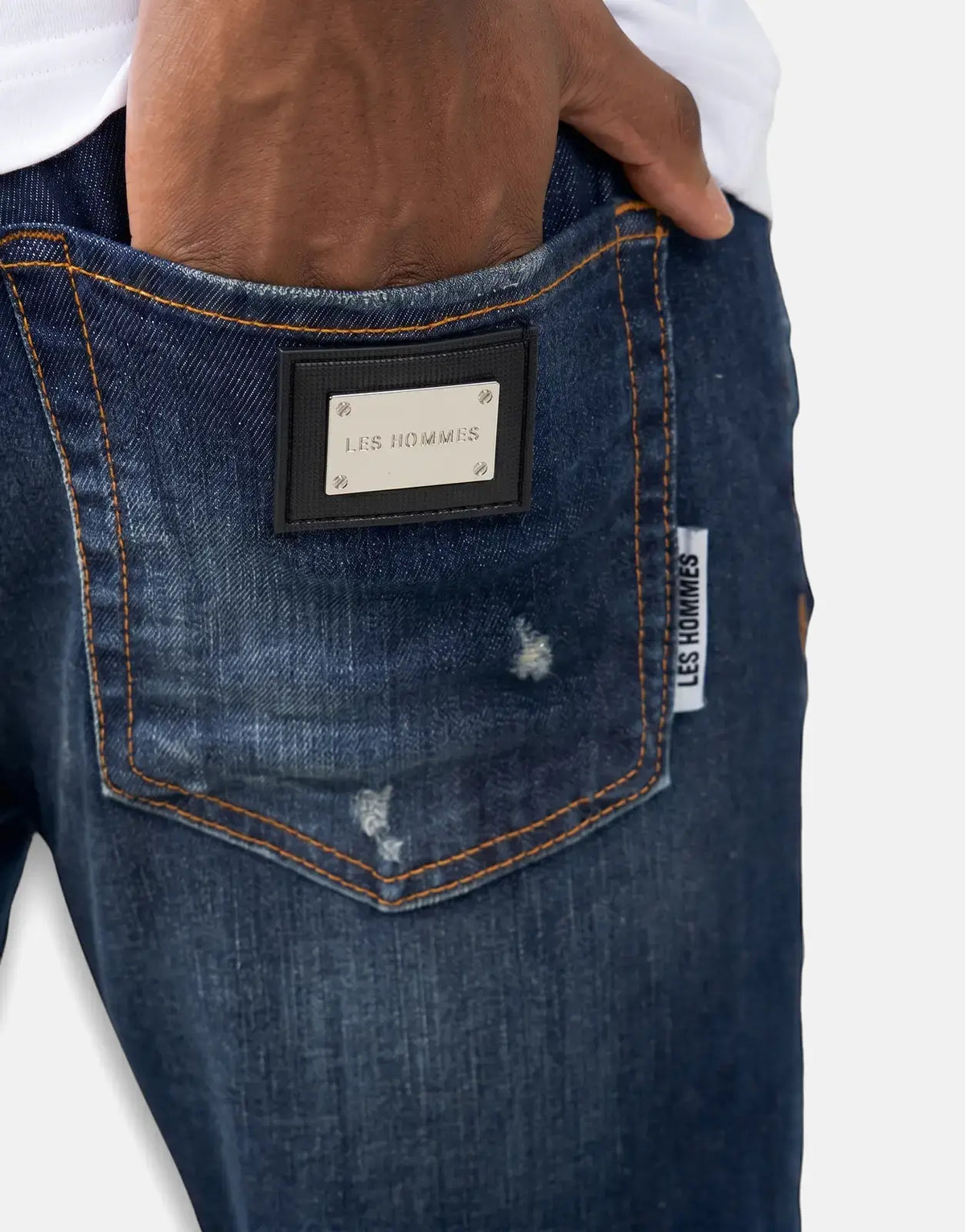Close-up of Les Hommes dark wash jeans with a hand in the back pocket, featuring a metallic brand patch and distressed