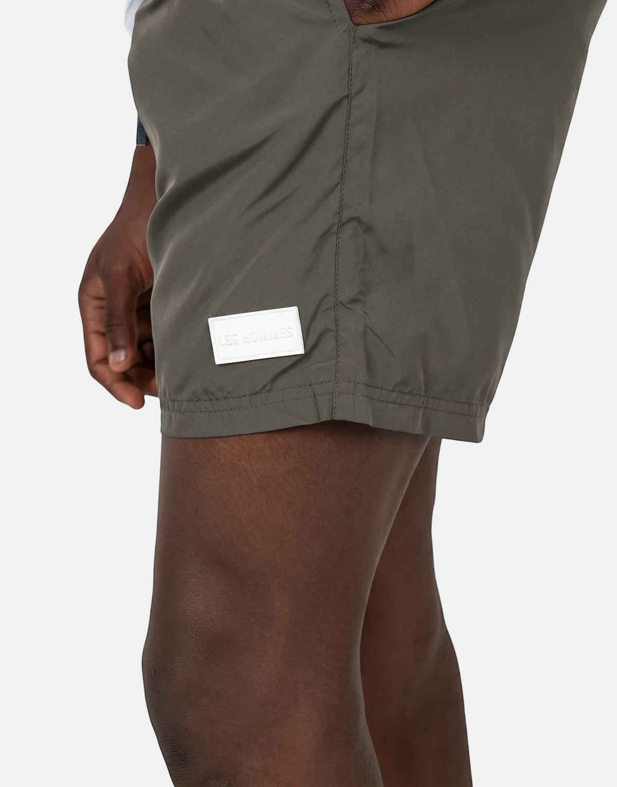 Close-up of Les Hommes olive green swim shorts worn by a model, featuring a white rectangular logo patch on the thigh.