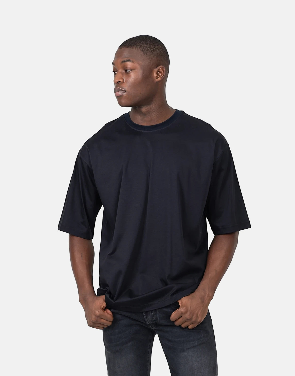 Young man wearing a black Les Hommes oversized t-shirt and dark jeans, hands in pockets, posing against a white background.