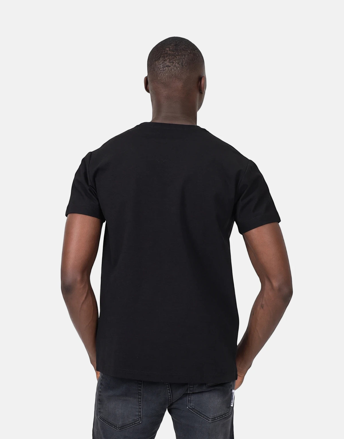 Back view of a Black man wearing a plain black Les Hommes t-shirt and dark grey jeans in a studio shot.