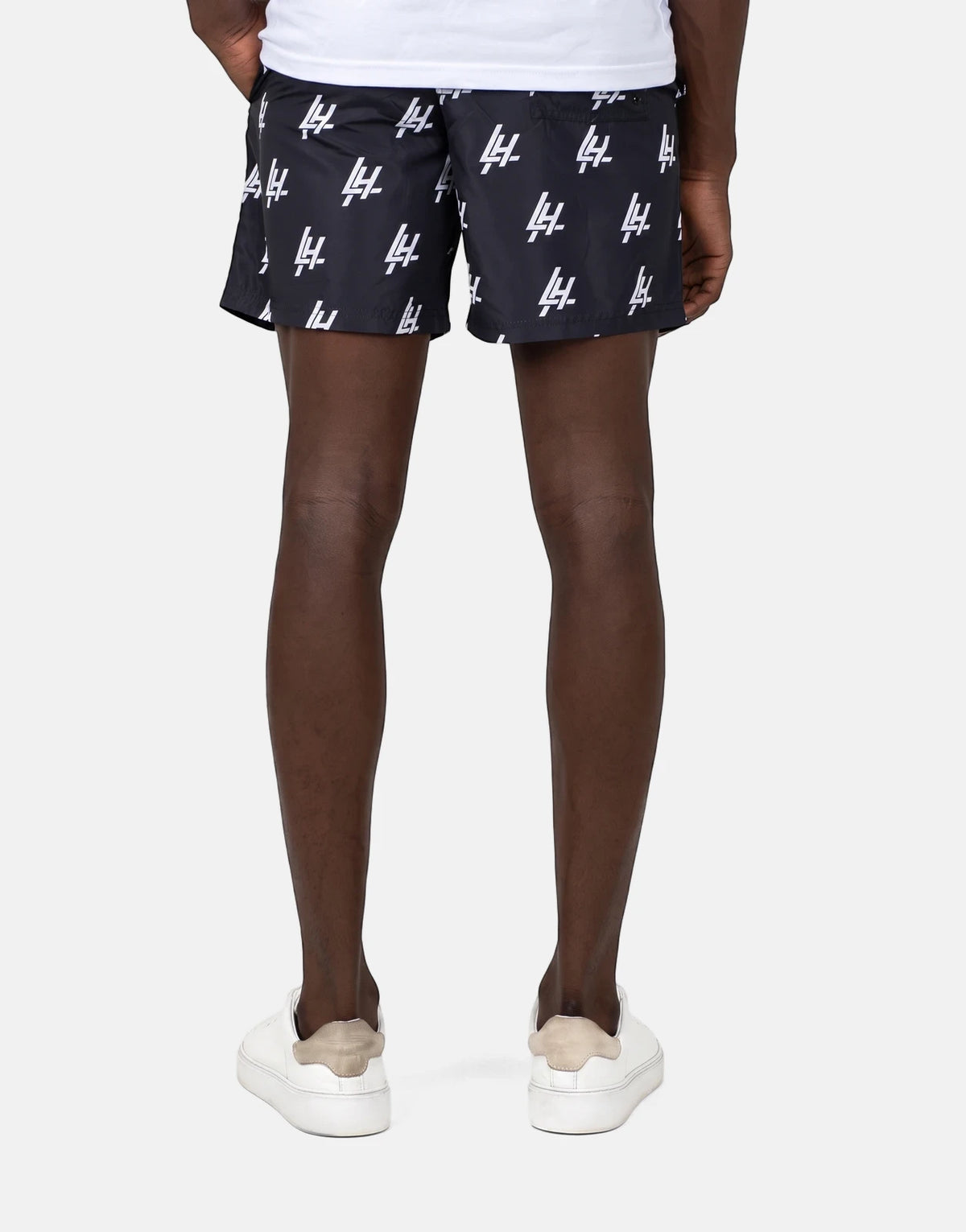 A person's lower body from behind, wearing black Les Hommes swim shorts with white logo print and white sneakers.