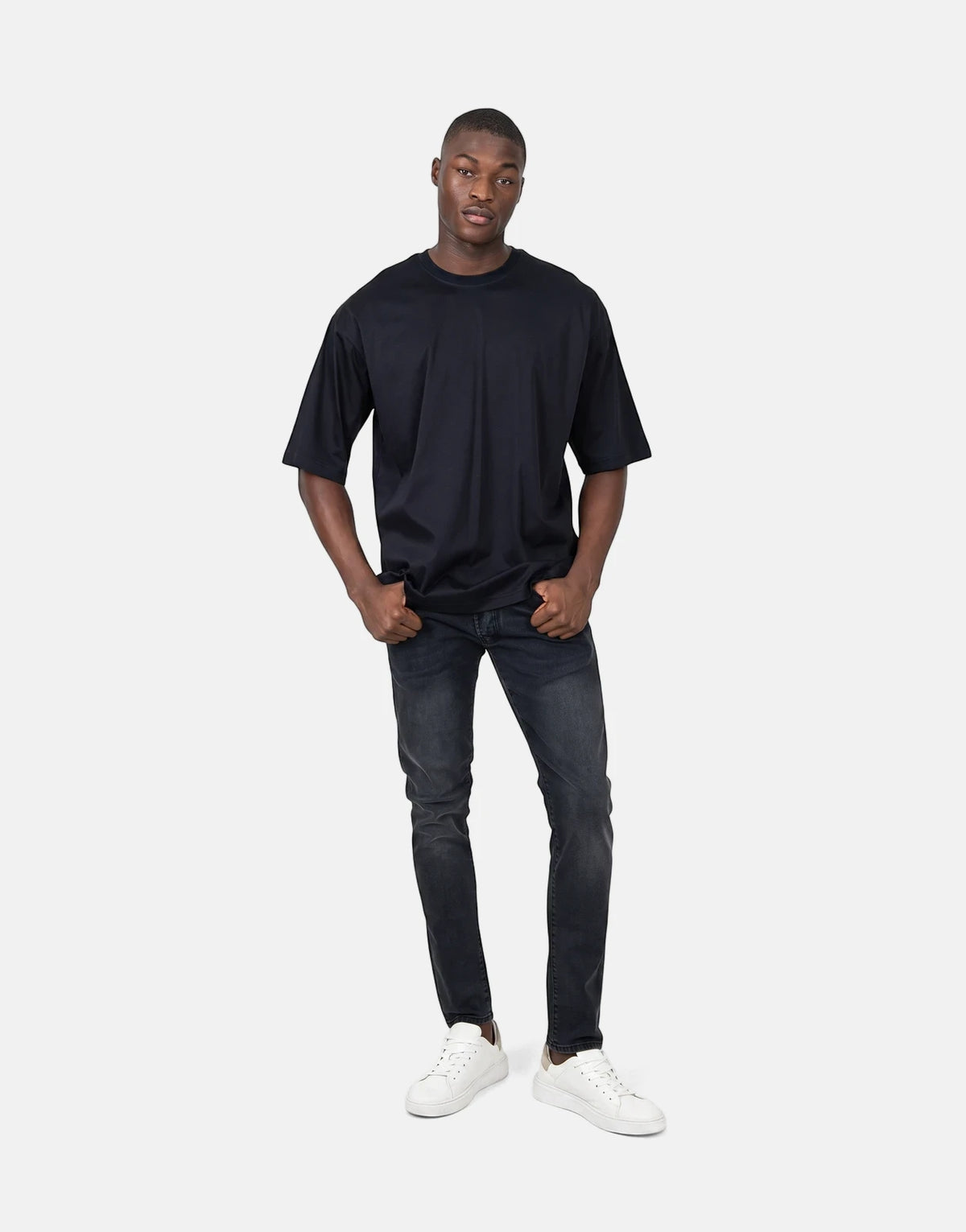 Male model wearing a black oversized t-shirt, black distressed jeans, and white sneakers, standing full body against a white