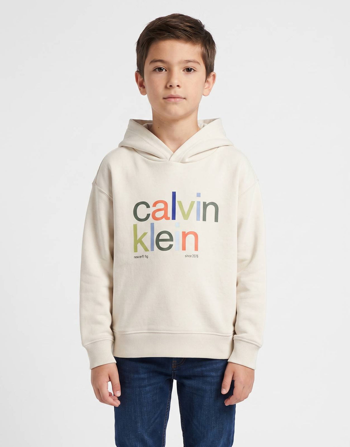A young boy wears a beige Calvin Klein Kids Color Logo Hoodie with blue denim jeans and white sneakers, shown in a full body