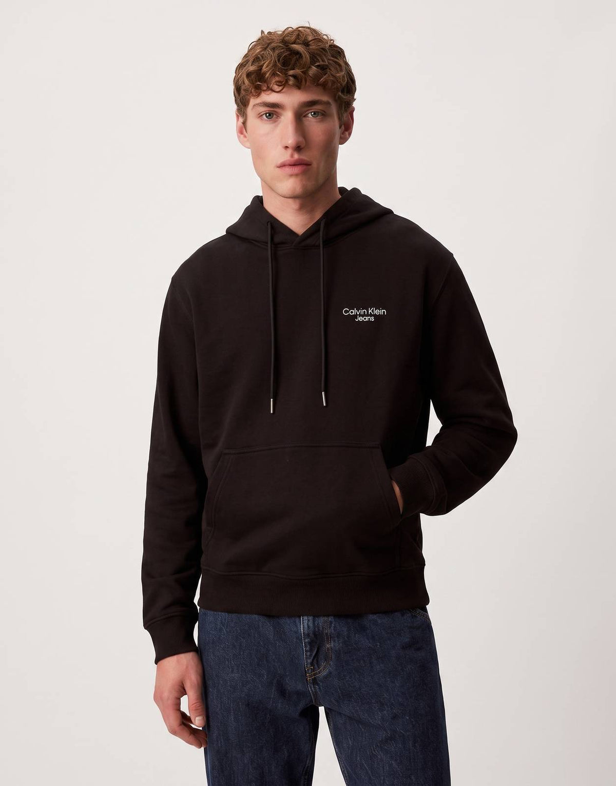 A male model wears a black Calvin Klein logo hoodie with a front pouch pocket, paired with dark jeans. Front view.