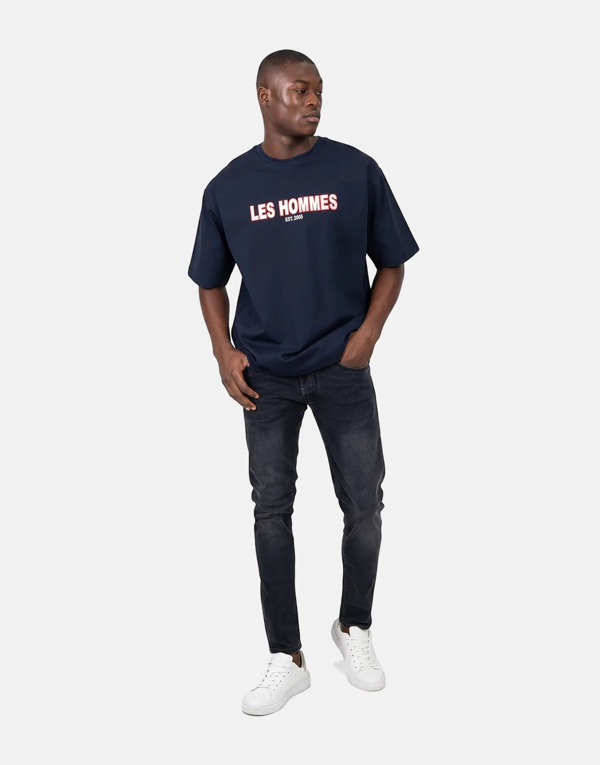Male model wearing a Les Hommes Over Rubber Print Blue T-Shirt with black jeans and white sneakers, full body shot.